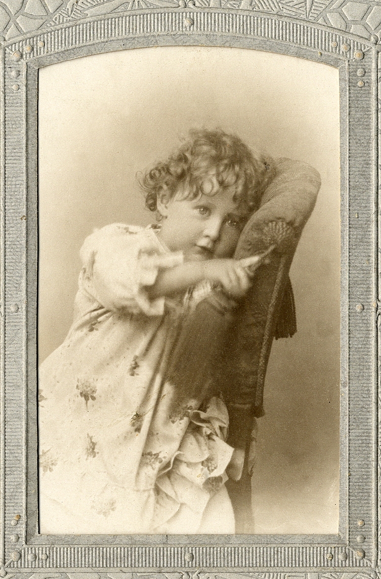 En liten flicka i småblommig klänning sitter på en stoppad stol, med en tofs på ryggstödet. 
Knäbild. Ateljéfoto.

Fotografens dotter.