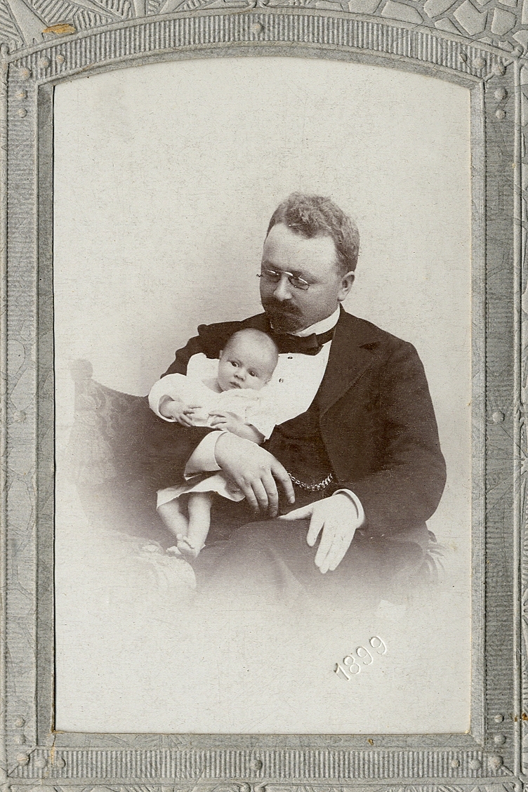 En man i mörk kavajkostym med väst, stärkkrage och fluga. Han håller en liten baby i famnen. I nedre högra hörnet syns inpräglat årtal: "1899". 
Knäbild. Ateljéfoto.
