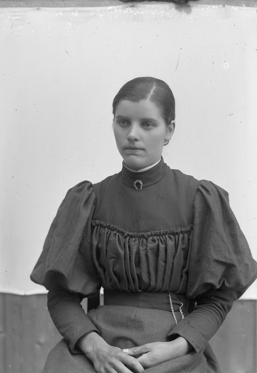 Kvinne med mørk kjole og kjede i beltet, hesteskonål v/halsen. Petra Brænna f. Nyhuus.