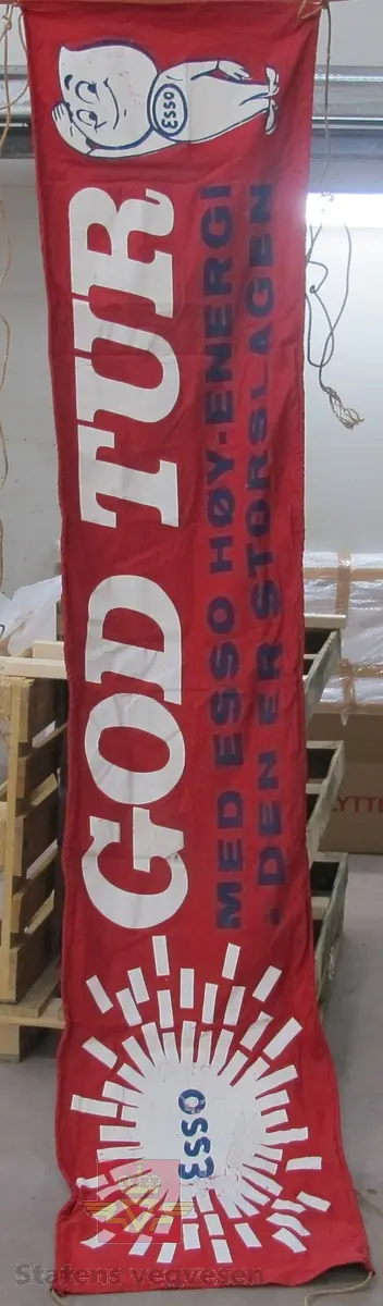Rød reklamebanner i tekstil med Esso logo og Dråpemannen. Har teksten: GOD TUR MED ESSO HØY-ENERGI - DEN ER STORSLAGEN. Banneren har langsgående sydde kanter der det er tredd inn tau til opphenging. Den er tosidig og har trykk på begge sider.
