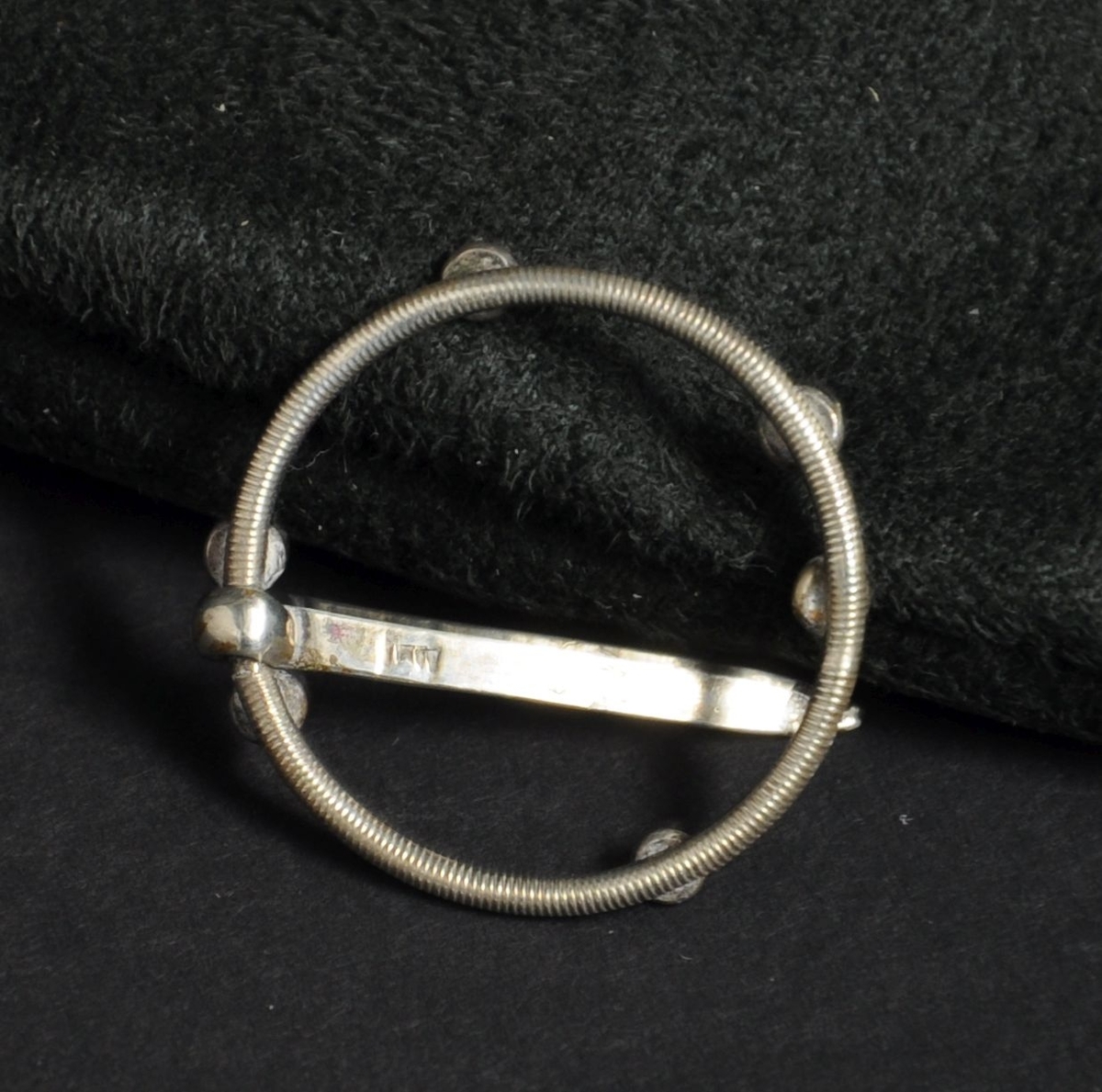 Spenne av sølv. Spennen har sirkulær form med dekorative tverrlinjer, og seks runde kuler.