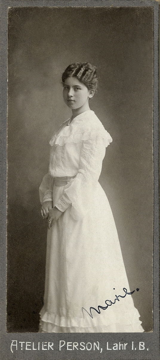 Porträttfoto av en ung kvinna i ljus klänning med volanger. 
I nedre högra hörnet syns autograf; "Marie".