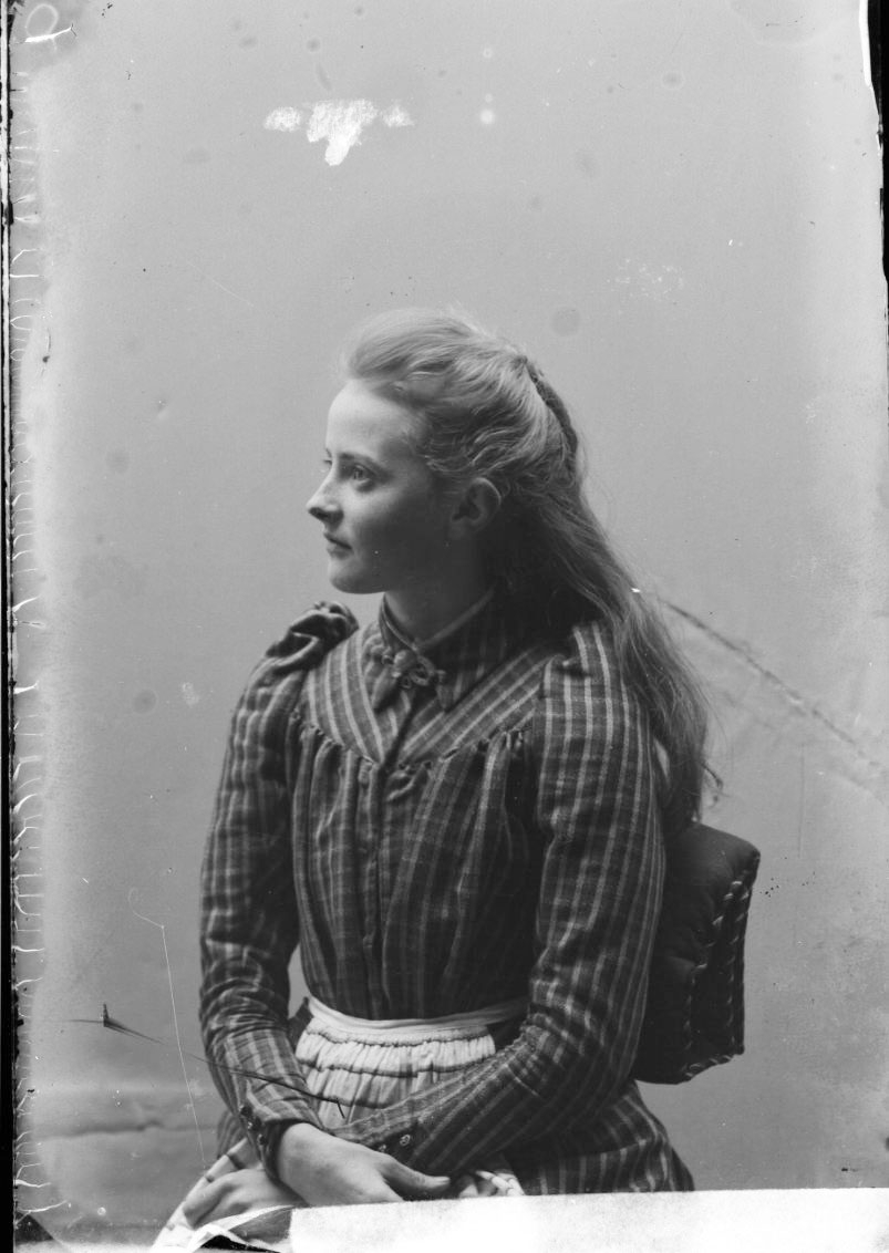 Porträtt av Ester Grönberg, sittande i profil. Hon sitter på en stol, klädd i rutig klänning och förkläde.