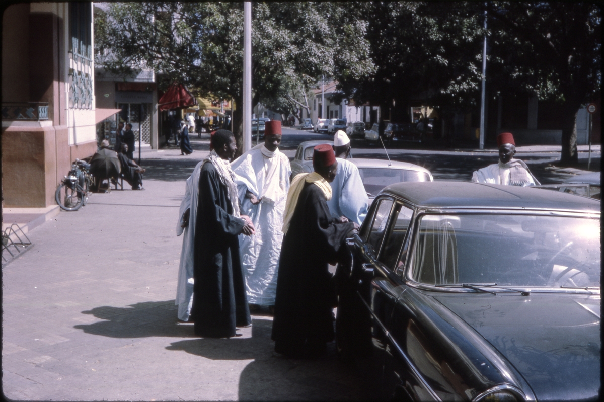 To svartkledde og fire hvitkledde menn, fire av dem iført fez, står ved parkerte biler. 'Sagafjord' Around The World via Africa Cruise 1966.
