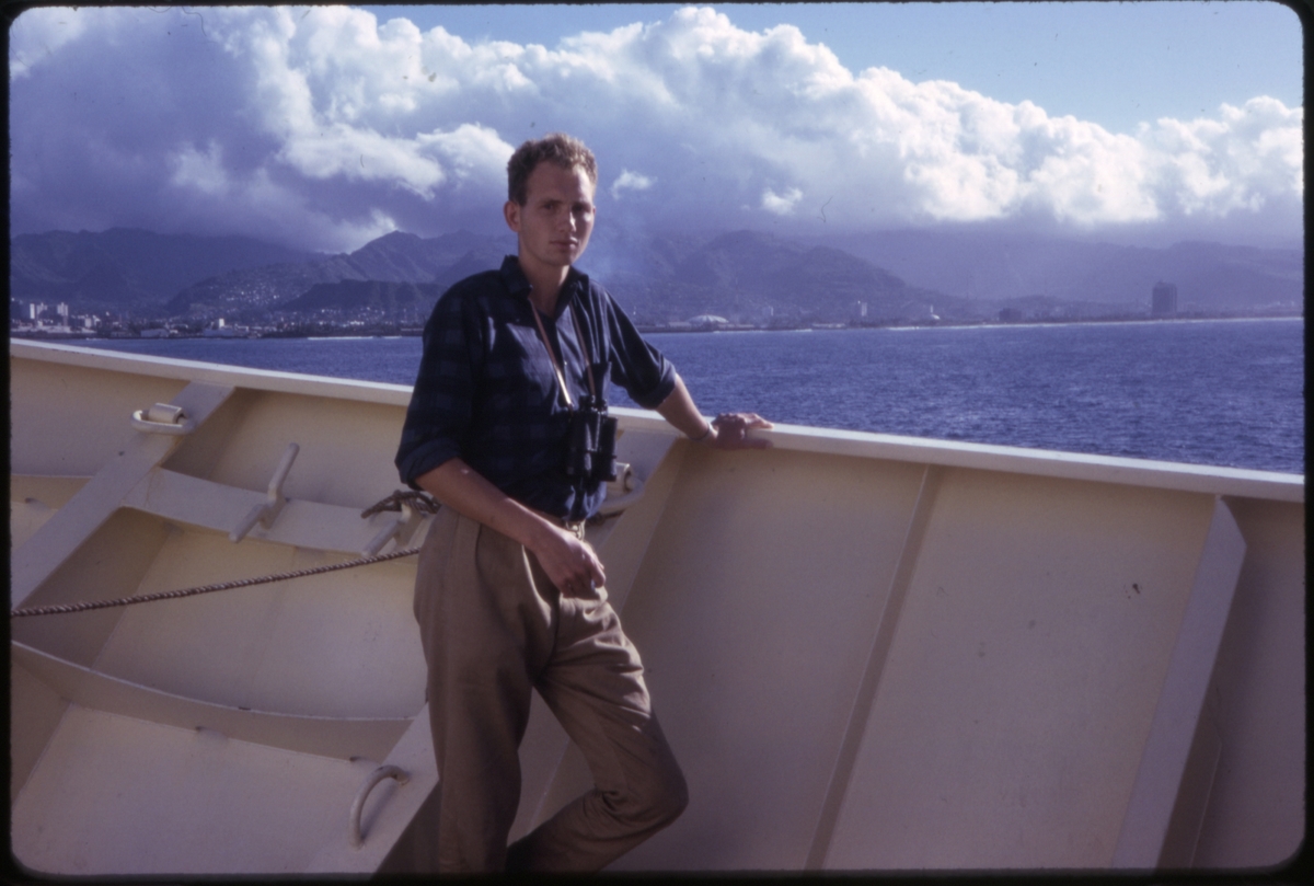 Mannlig turist i baugen på cruiseskipet 'M/S Sagafjord', med skyer, fjell og bebyggelse i bakgrunnen, trolig ved Hawaii. 'Sagafjord' Around The World via Africa Cruise 1966.
