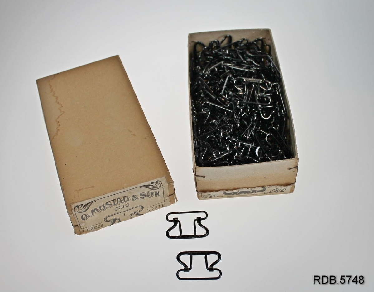 4 pappesker med lokk som inneholder svarte metallhemper fra O. Mustad & sønn. Hempene har to tagger til å stikke ned i stoffet den skal være på.