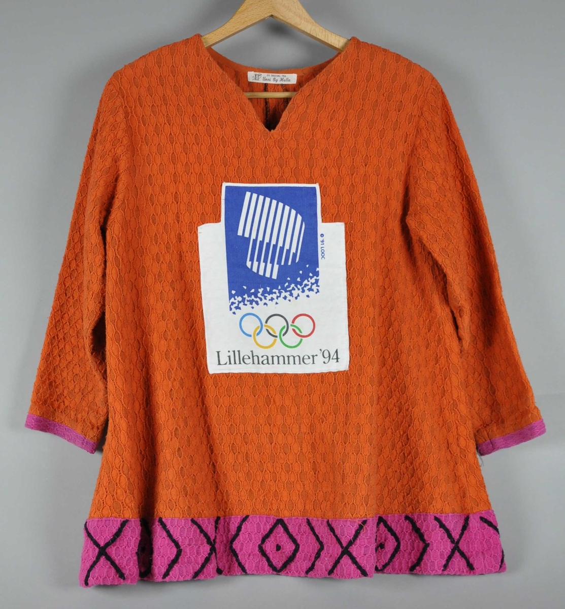 Oransje trøye av vevet bomull med emblem for Lillehammer '94 på brystet. Trøya  har v-hals og en rosa kant med svart brodering nederst og rosa kanter  ytterst på begge ermer.