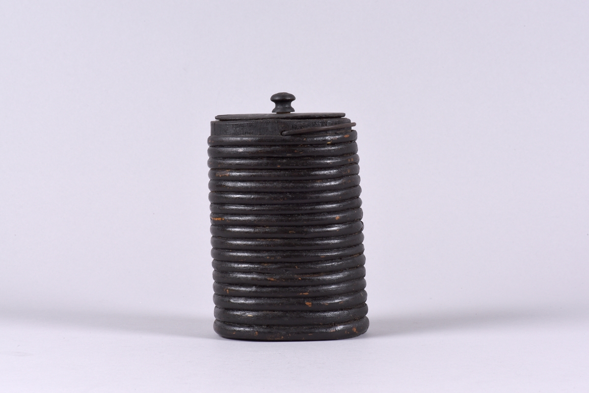 Oval, laggad brännvinsflaska av trä med täta band. Lock med svarvad knopp och litet uttag. Svartmålad, troligen sekundärt.