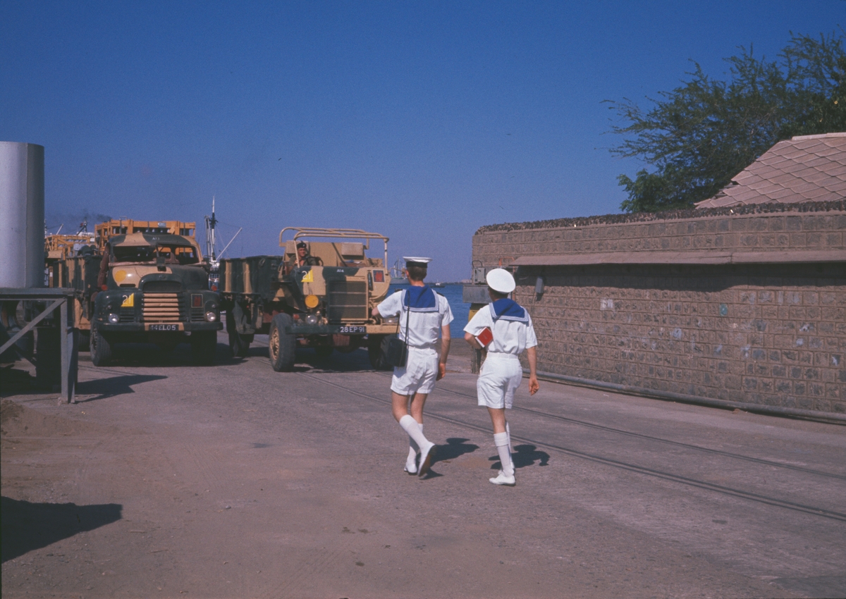 Bilden visar två svenska sjömän i sjömanskostym som promenerar i Aden. I bakgrunden syns två engelska militär lastbilar.