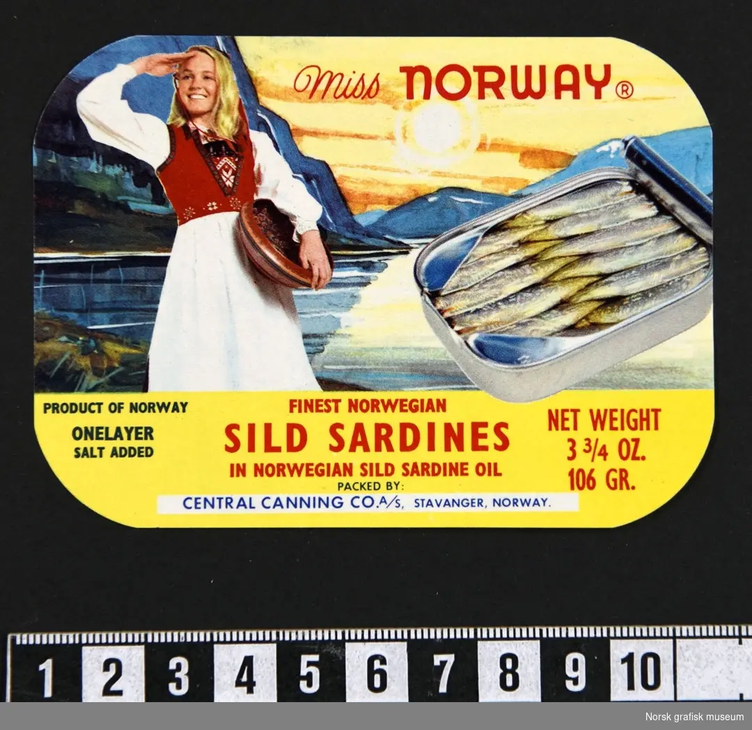 Etikett med en blond speidende kvinne i bunad foran et malerisk landskapsmotiv. Ved siden av er det bilde av en åpnet hermetikkboks. 

"Finest Norwegian sild sardines in Norwegian sild sardin oil"