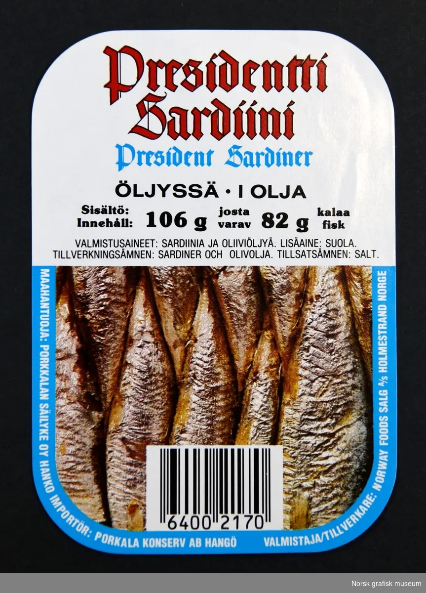 Etikett med hvit bakgrunnog detlajer i lys blå og rødt. Halve etiketten er dekt av bilde av innholdet i boksen: 
sardiner i olje.