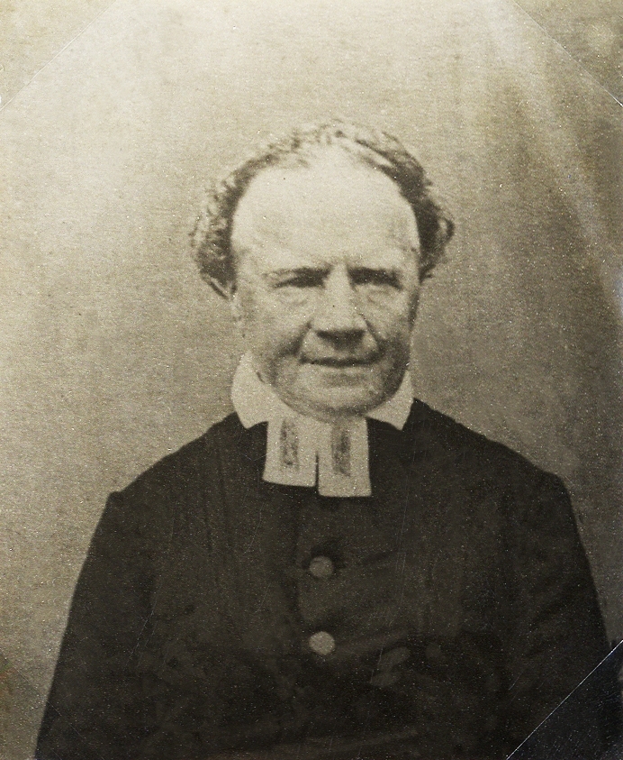 Foto av en äldre man i prästrock med prästkrage m.m.
Bröstbild, halvprofil. Ateljéfoto.