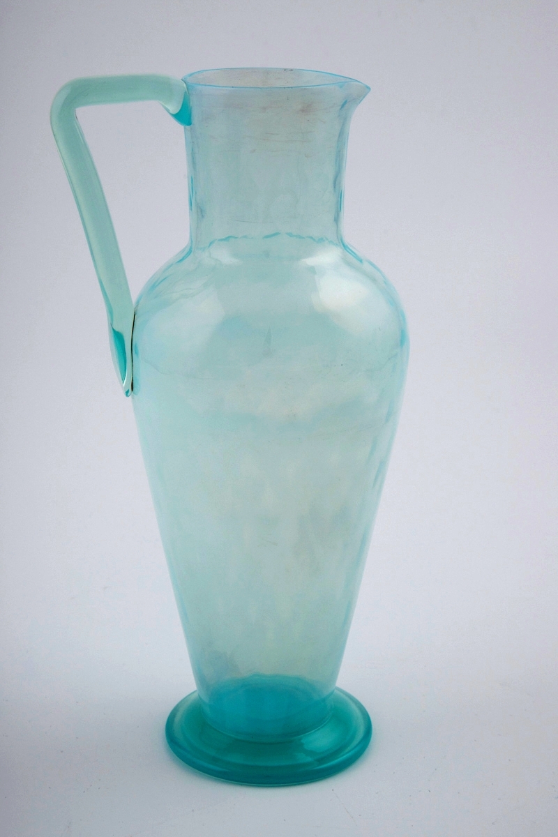 Høyreist mugge med hank i blålig opaliserende glass. Nedre del av muggen er tilnærmest opak, glasset blir gradvis mer gjennomskinnelig mot øvre del av korpus og den korte halsen. Dråpeformede rifler på korpus og hals.