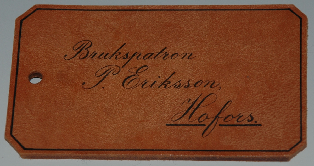 Adresslapp i brunt läder, 13,3 x 7,6 x 0,4 cm.
Rektangulär med fasade hörn.
Ram och text i svart: "Brukspatron P. Eriksson, Hofors"