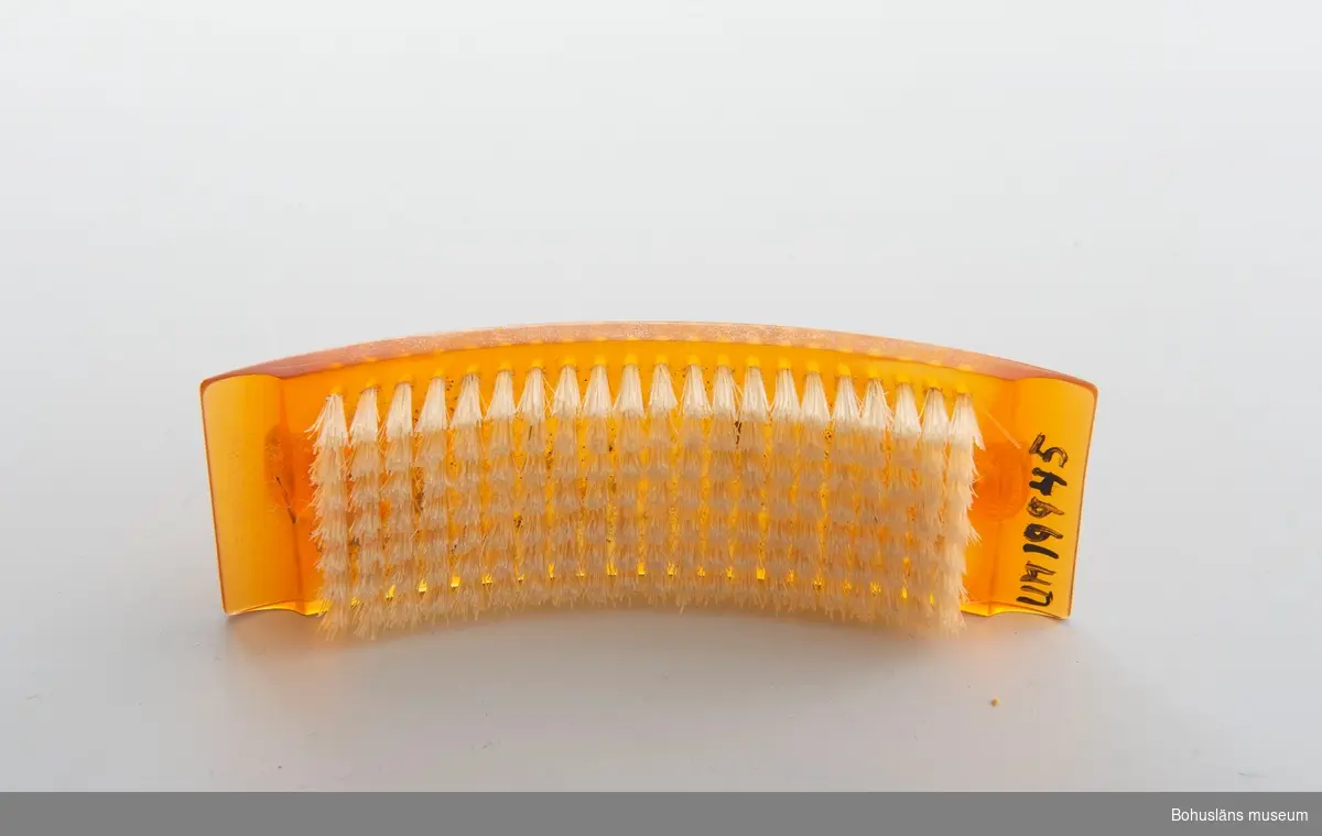 Konkav nagelborste i genomskinlig orange plast med bågformat handtag. 
Försedd med nylonborst.