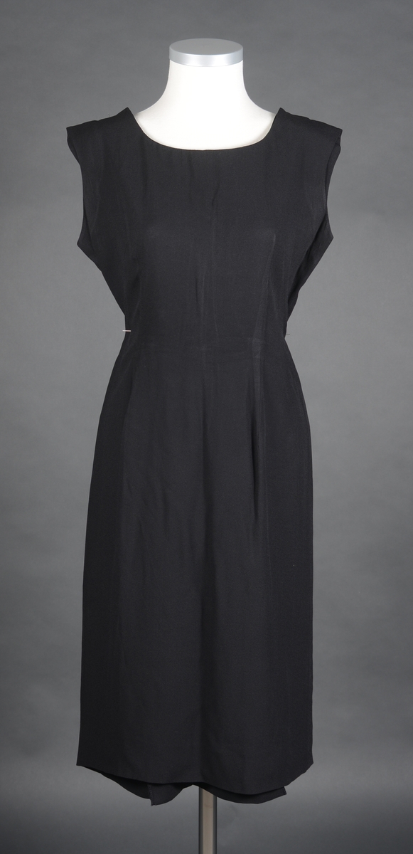Ermeløs kjole sydd av svart kunstsilke. Den har enkel utforming. Midt bak er det en splitt. Kjolen hører sammen med jakke FTT.33011.