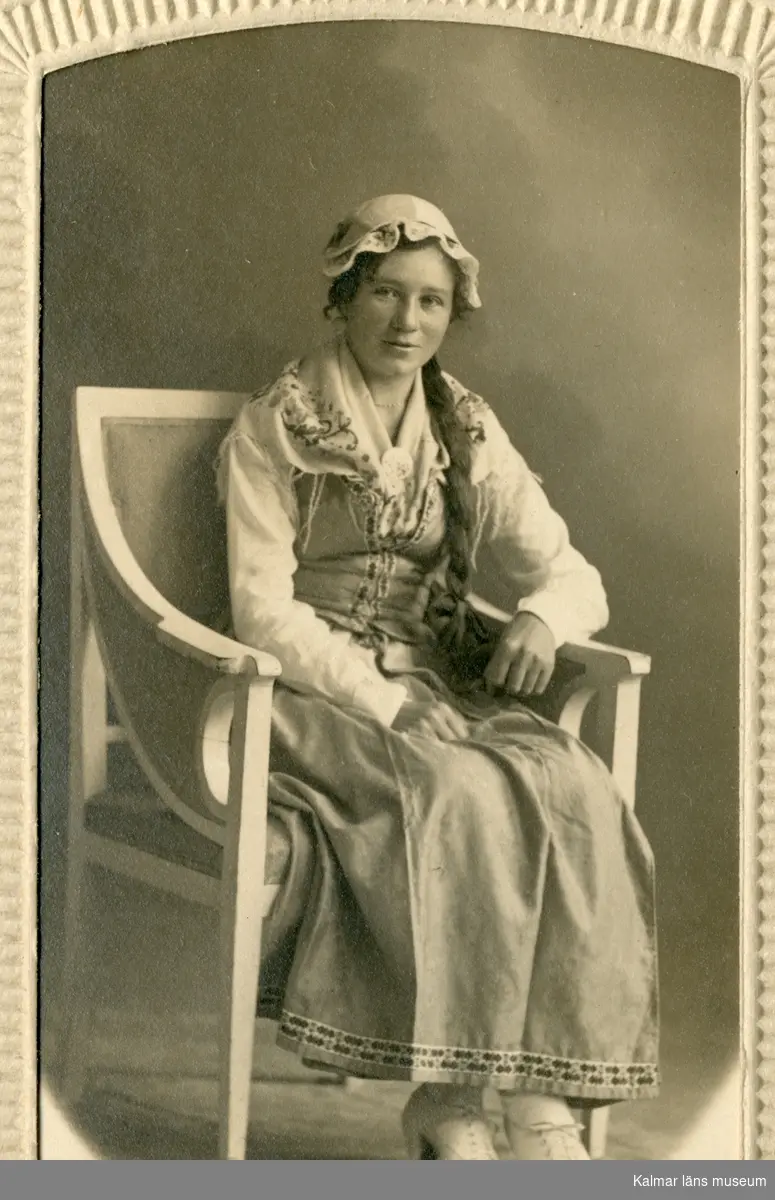 Porträtt av Esther Sivgård, klädd i hätta, sjal och klänning sitter i en karmstol.