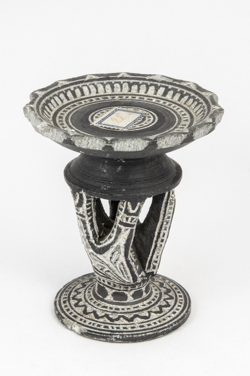 Litet bord av sten med tre ben och fotställning samt ornament, kanten krusig, från Mekka . 

Avd. Österländska föremål i Ahléns tryckta katalog.