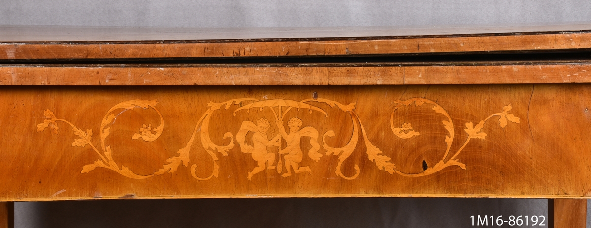 Spelbord av mahogny. På framsidan av sargen dekorationer i intarsia. Skivan, nedfällbar, är liksidig med avrundade hörn. Raka, nedåt avsmalnande ben. Se 1M16-86191.