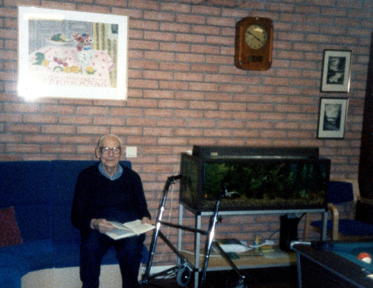 En okänd man sitter i en soffa och bläddrar i en bok, Brattåsgården (Streteredsvägen 5) cirka 1990. Vid sidan av mannnen står hans rullator samt ett akvarium. Bakom mannen är en gul tegelvägg.
