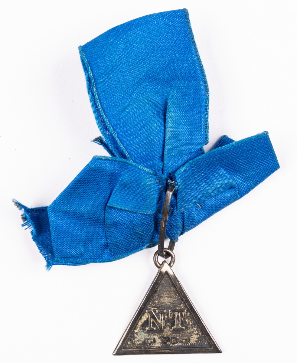 Triangelformat hänge i blå sidenrosett av siden.
Sällskapet NT. Stämplad.