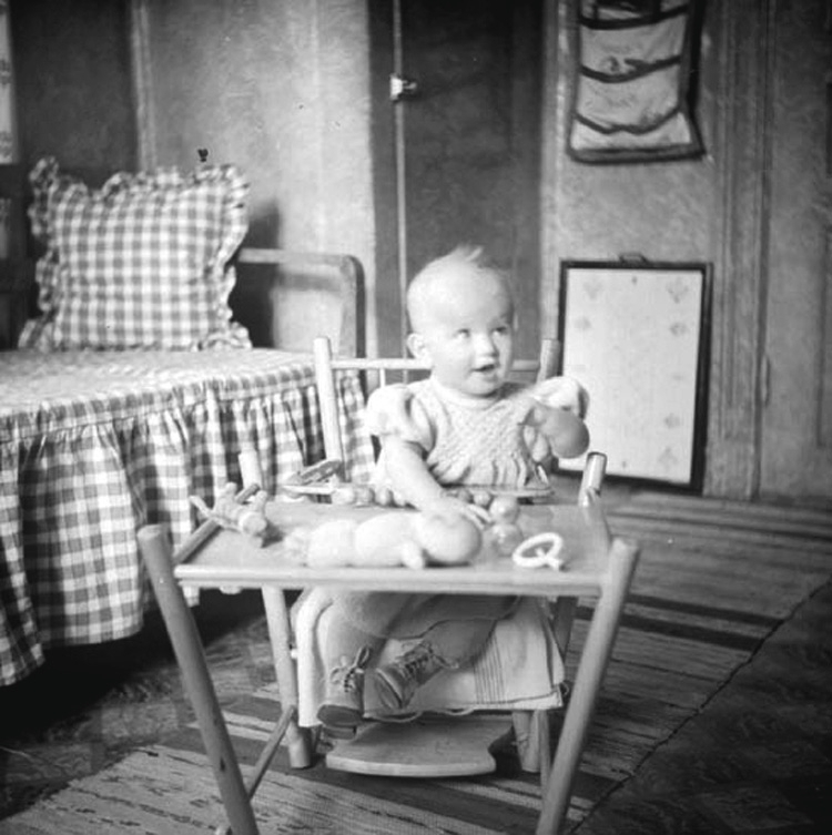 Ett litet barn sitter i en hopfällbar barnstol av trä med leksaker framför sig.