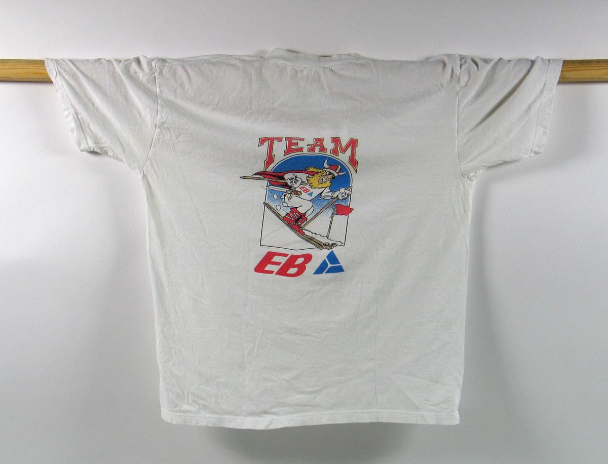 Hvit t-skjorte. Foran er det en logo for de olympiske vinterleker på Lillehammer i 1994. På ryggen er det en logo for TEAM EB. I denne logoen inngår det et motiv av en viking på ski.