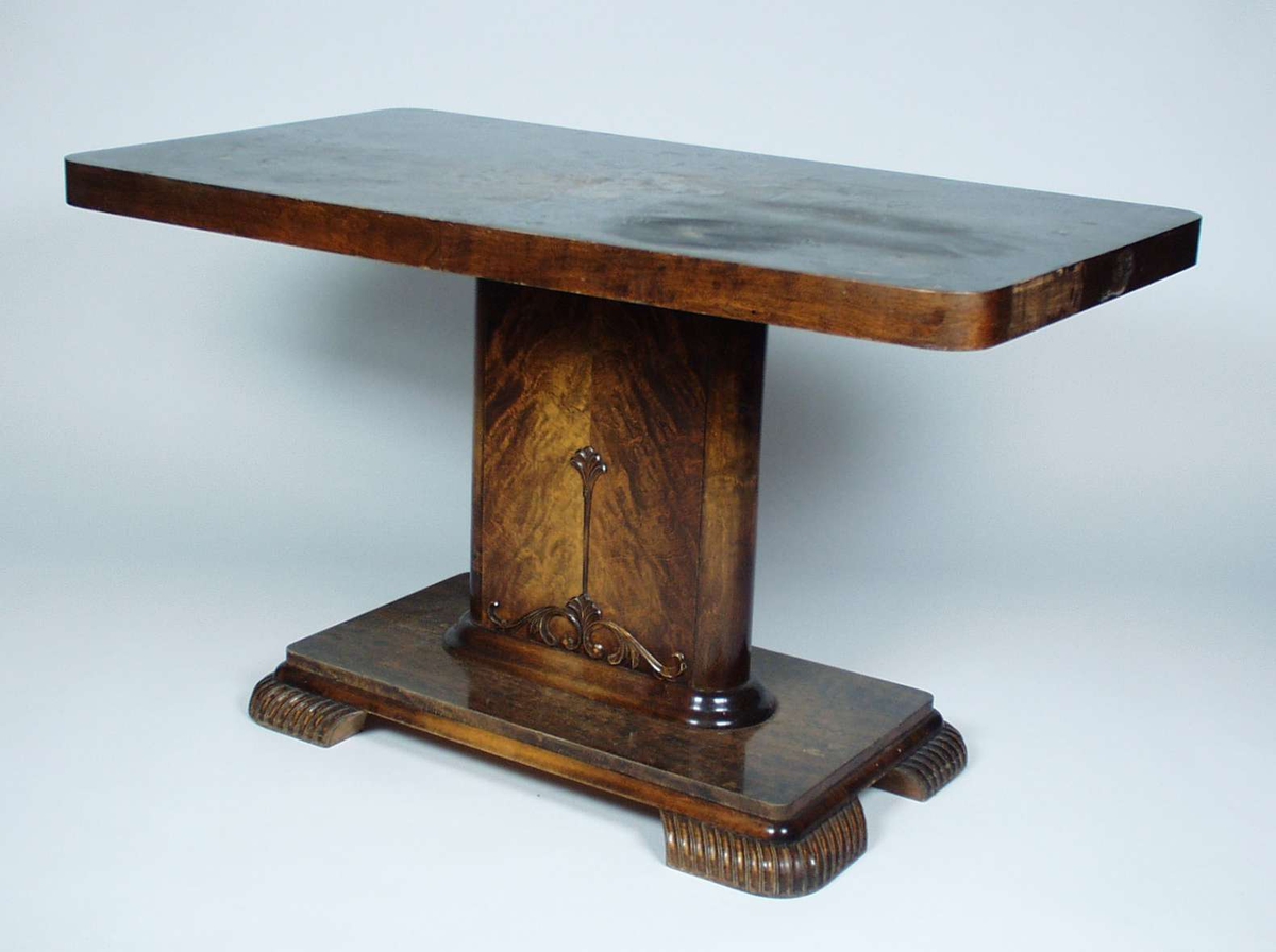 Et bord i furu som er lakkert i brunfarge.
Det har firkantet midtsøyle med utskjæring i form av blomst. Midtsøylen står på en plate med fire ben.

