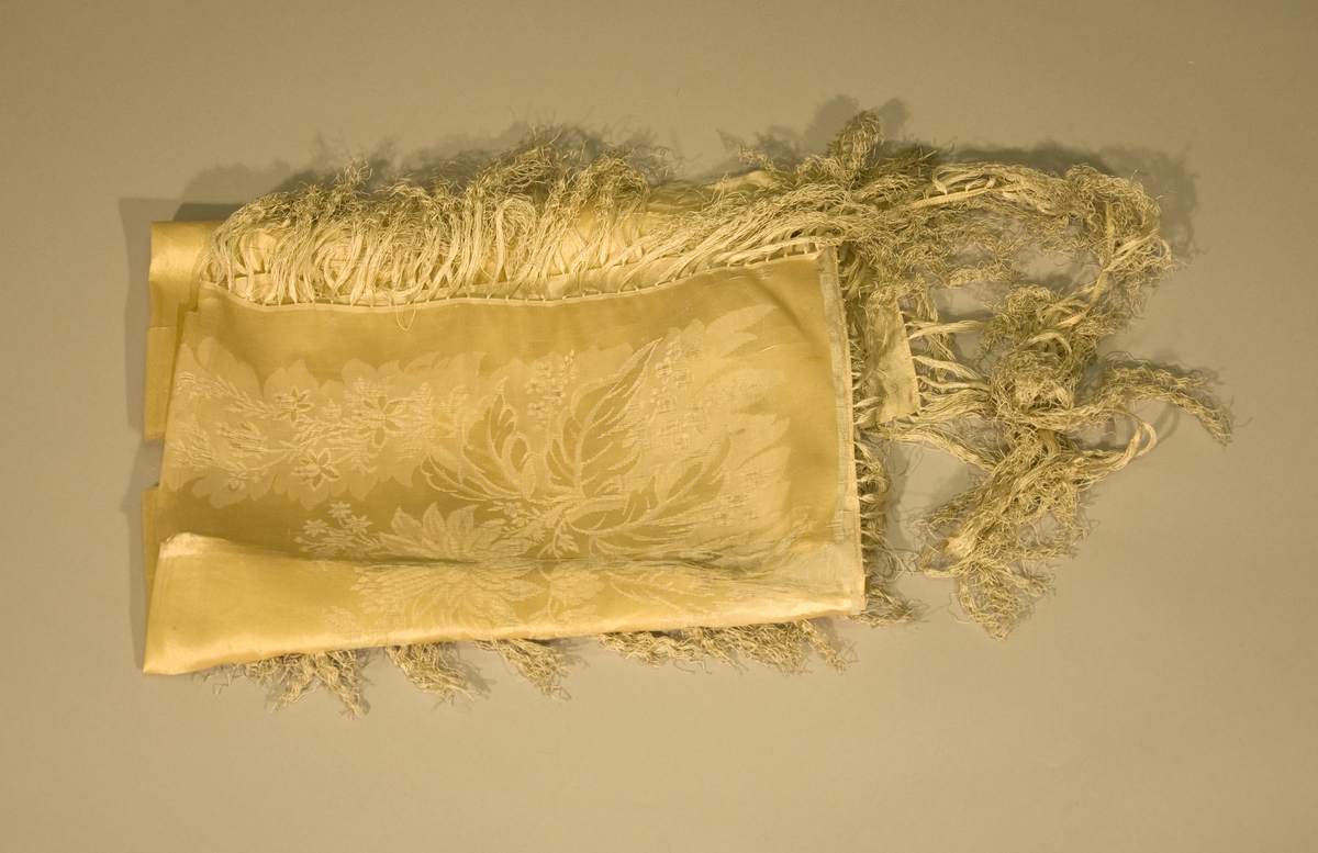 Gult tørkle av mønstervevd silke. Tørkledet har maskinsydd fald og er påsydd silkefrynser, 11 cm. I svært dårlig stand, falden og frynsekanten er delvis løsnet fra tørkledet.