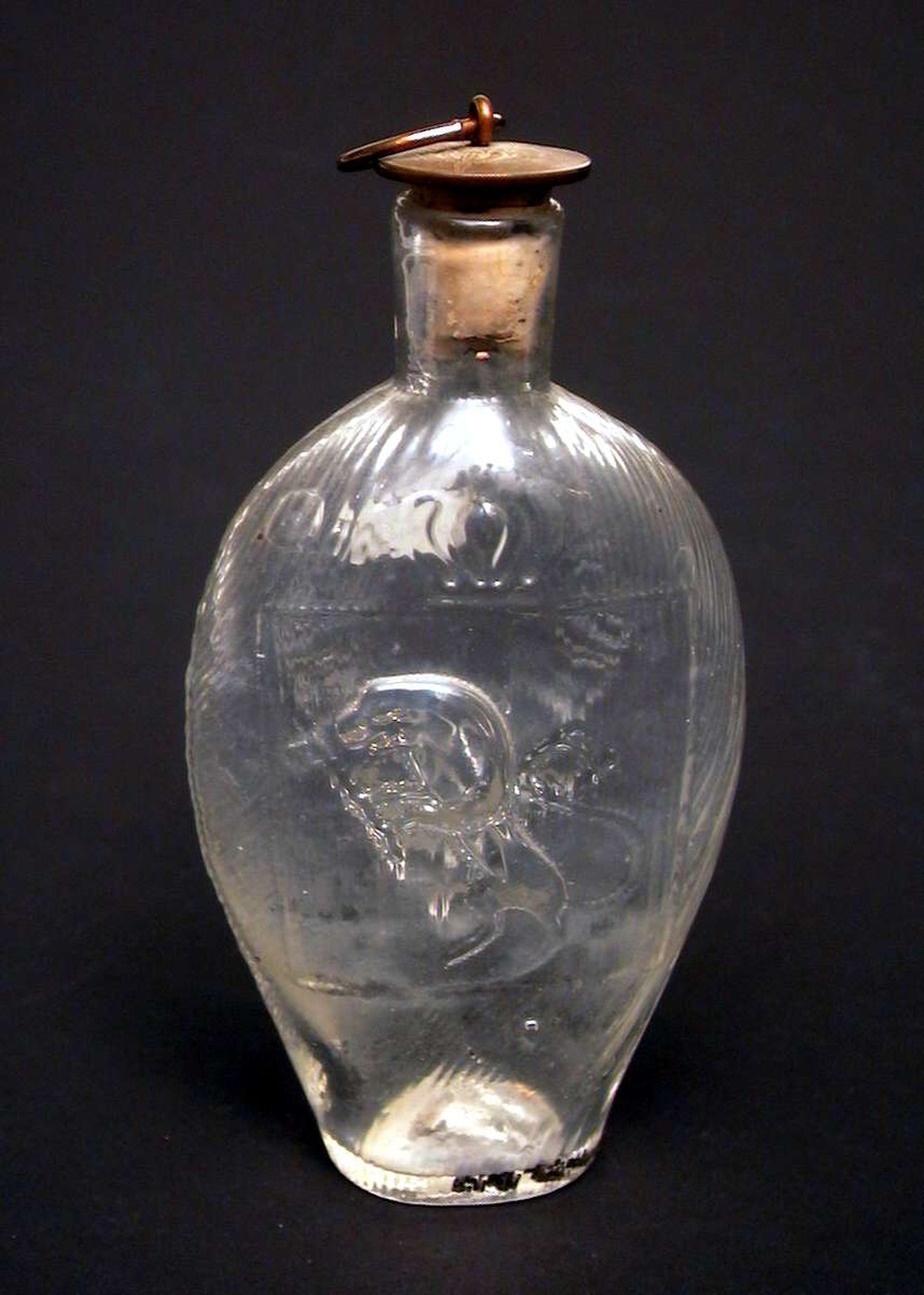 Jaktflaske med buet, flat korpus. Den er dekorert med en drueklase på den ene siden, på den andre en løve. Flasken har kork med metallbeslag. På beslaget står det 'Gustaf VI Adolf Sveriges Konung'.