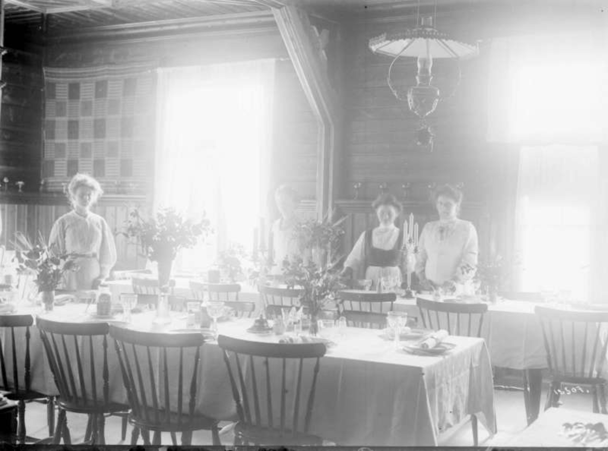 03.08.1909. Kvam hotel. Interiør av spisestuen, dekket og blomsterpyntet bord, stoler, veggteppe, gardiner, taklampe, tre kvinner.