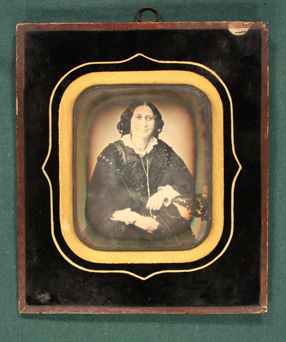 Portrett av Kaja Westergaard, 1856. Portrett av kvinne i en face, sittende, avsk. ved knærne, langt sort hår  med midtskill. Hvit blondekrave med brosje, holder med begge hender om en lang  lenke. Sort omramn. med gull inneramme.