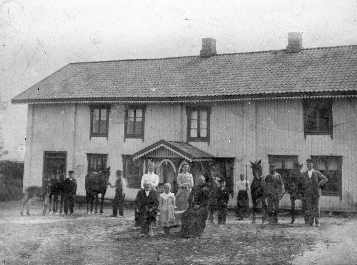 Nitteberg. Familen, tjenestefolk og hester samlet på tunet foran hovedhuset. Trolig i slåtten, ei jente i bakgrunnen står med ei rive.