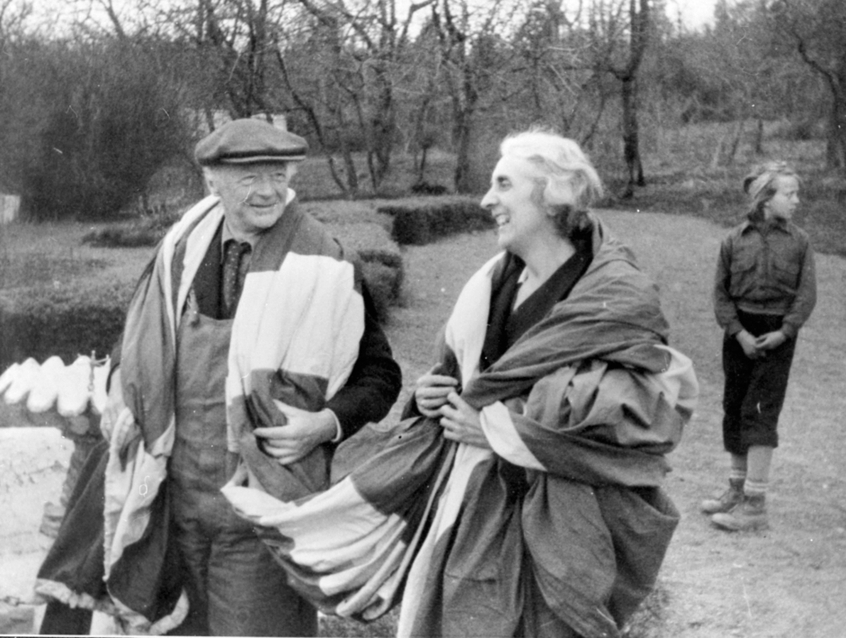 Domprost Reidar Kobro og bispinne Julie Krohn-Hansen med det norske flagg over skuldrene på Hovelsrud, Helgøya i fredsdagene i mai 1945. Nora Johanne Hagen f. 1933 (gift Rise) i bakgrunnen.