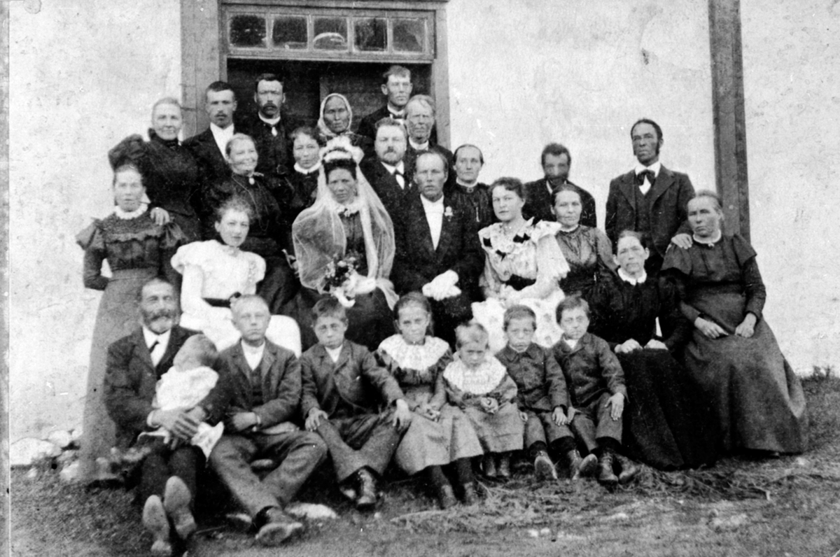 Brudepar og stor gruppe bryllupsgjester på Fjell gård, Helgøya. Brudeparet er Otlie Torsdatter (1855-1927) og Kristian Bratberg (1860-1932). Foran hovedbygningen på Fjell. Foran er noen av barne på Nedre Maurud.