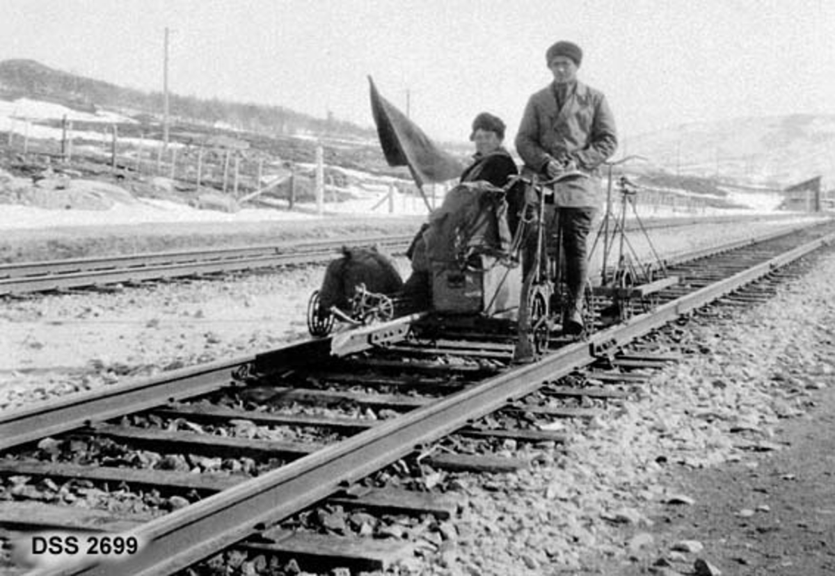To mann på dresin ved Vålåsjø stasjon på Dovrefjell.  En mann sitter på sykkel, den andre er passasjer med redskapskasse og flagg.  Bildet er tatt på dobbeltsporet strekning.  Fjellandskap i bakgrunnen. 