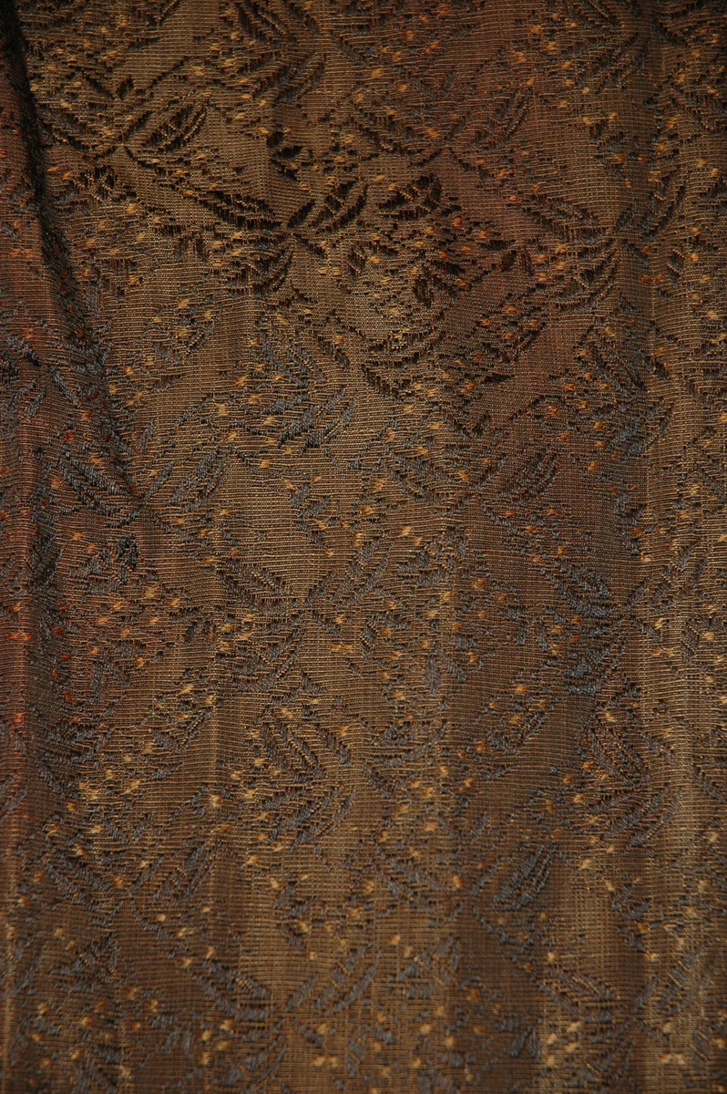 Langt, brunt skjørt med mønster i lysebrunt og svart. Skjørtet er foret med mørk brunt bomullsstoff og har kantbånd nederst i mørk brunt. Rynket parti midt bak. Lomme i høyre side.