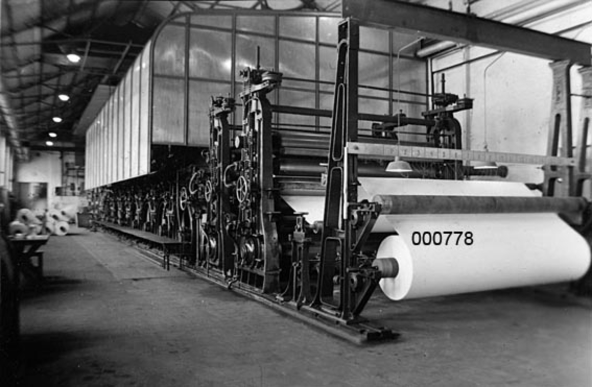Interiør fra A/S Buskerud Papirfabrikk på Åssida i Drammen (den gang i Lier kommune).  I 1937-38 ble papirmaskinen ved denne fabrikken fullstendig ombygd, særlig for å kunne kjøre maskinen med større driftshastigheter, slik at en kunne oppnå større produksjonsvolumer.  I den forbindelse la bedriften om fra dampdrift til elektrisk drift. Dette fotografiet viser maskinen sett fra opprullingsenheten i enden av maskinsalen.  Bakenfor ses glitten og det overbygde tørkepartiet.  Ombyggingsarbeidet ble utført av A/S Thunes mekaniske Værksted i Oslo i samarbeid med Siemens. 