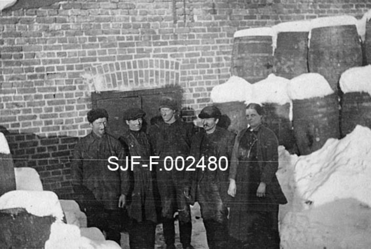 Fem ansatte ved Schwencke & Co's bek- og tjærefabrikk ved Kongshavn sør for Oslo, fotografert foran en av inngangene til fabrikklokalet en kald vinterdag, antakelig omkring 1. verdenskrig.  Vi ser (fra venstre) fire menn og ei kvinne.  Mennene har arbeidstøy (overaller) og hodeplagg, kvinna en mørk kjole, samt ei jakke på overkroppen.  Bak dem ser vi en teglsteinsvegg med ei dobbelt inngangsdør.  På begge sider av døra er det stablet tretønner på høykant, tønner som antakelig inneholdt tjære eller bek.  Så vel tønnene som terrenget omkring dørene preges av betydelige snømengder. 