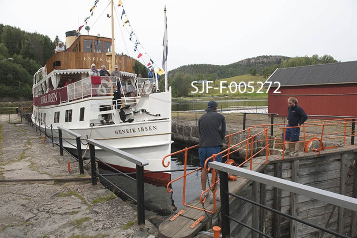Turistbåten «Henrik Ibsen» i Kjeldal sluse i Bandak-Norsjø-kanalen.  Sluseanlegget ligger i Nome kommune.  Her er det ett slusekammer, som utlikner en høydeforskjell på tre meter mellom ovenforliggende og nedenforliggende del av vassdraget.  Slusene betjenes i 2012 av to slusevoktere som reiser foran rutebåtene i bil.  Her ser vi karene på brua over nedre sluseport. Båten går i rute mellom Skien by og Dalen i Tokke, innerst ved Bandakvatnet, en distanse på 105 kilometer.  Henrik Ibsen er det største, raskeste og mest luksuriøse skipet i Skiensvassdraget.  Fartøyet er opprinnelig bygd som dampskip ved Eriksbergs Mekaniska Verkstad i Gøteborg i 1907 for Styrsö-bolaget.  Derfor fikk båten først navnet DS «Styrsö».  Under dette navnet gikk fartøyet i mange år som rutebåt i Gøteborg-skjærgården.  Etter en periode som charterbåt på den svenske vestkysten ble Styrsö oppkjøpt av Skien Dalen Skipsselskap AS og satt trafikk i Skiensvassdraget i forbindelse med Bandak-Norsjø-kanalens 100-årsjubileum i 1992.  Her fikk båten navnet «MS Henrik Ibsen», etter den berømte forfatteren med tilknytning til Skien og en hurtiggående dampbåt som gikk i vassdraget tidig på 1900-tallet.  I perioden 2009-2011 ble skipet restaurert med sikte på at det igjen skulle framstå som det hadde gjort da det var nytt.  Etter dette har fartøyet vært driftet av Norwegian Hospitality Group AS, som eier Telemarkskanalen Skipsselskap AS.  Selskapet disponerer også det tradisjonsrike drakestilhotellet på Dalen, som er endestasjon for båtruta.  Sesongen for kanalbåtene starter vanligvis i midten av mai og varte til andre halvdel av september. Seilasen mellom Skien og Dalen med turistbåten Henrik Ibsen tar cirka ni timer. MS Henrik Ibsen har aller rettigheter og det er restaurant ombord med daglig servering av frokost og lunsjretter.  Båten går opp den ene dagen og ned igjen den neste.  Turistbåten MS Victoria går ned til Skien den dagen Henrik Ibsen setter kurs for Dalen, og går ned igjen den dagen den andre båten er på veg opp. 