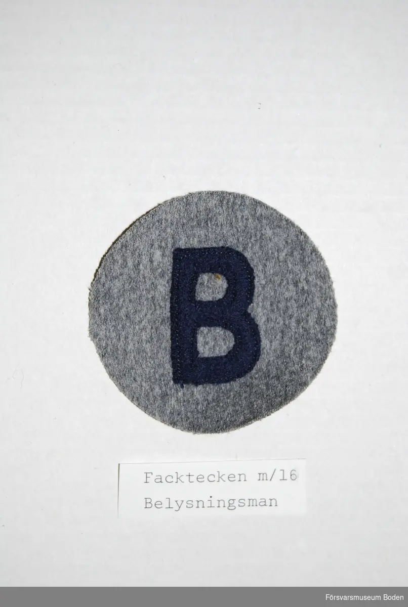 Bokstaven B av mörkblått kläde på grå bakgrund. Syddes fast på uniformens vänstra underarm.