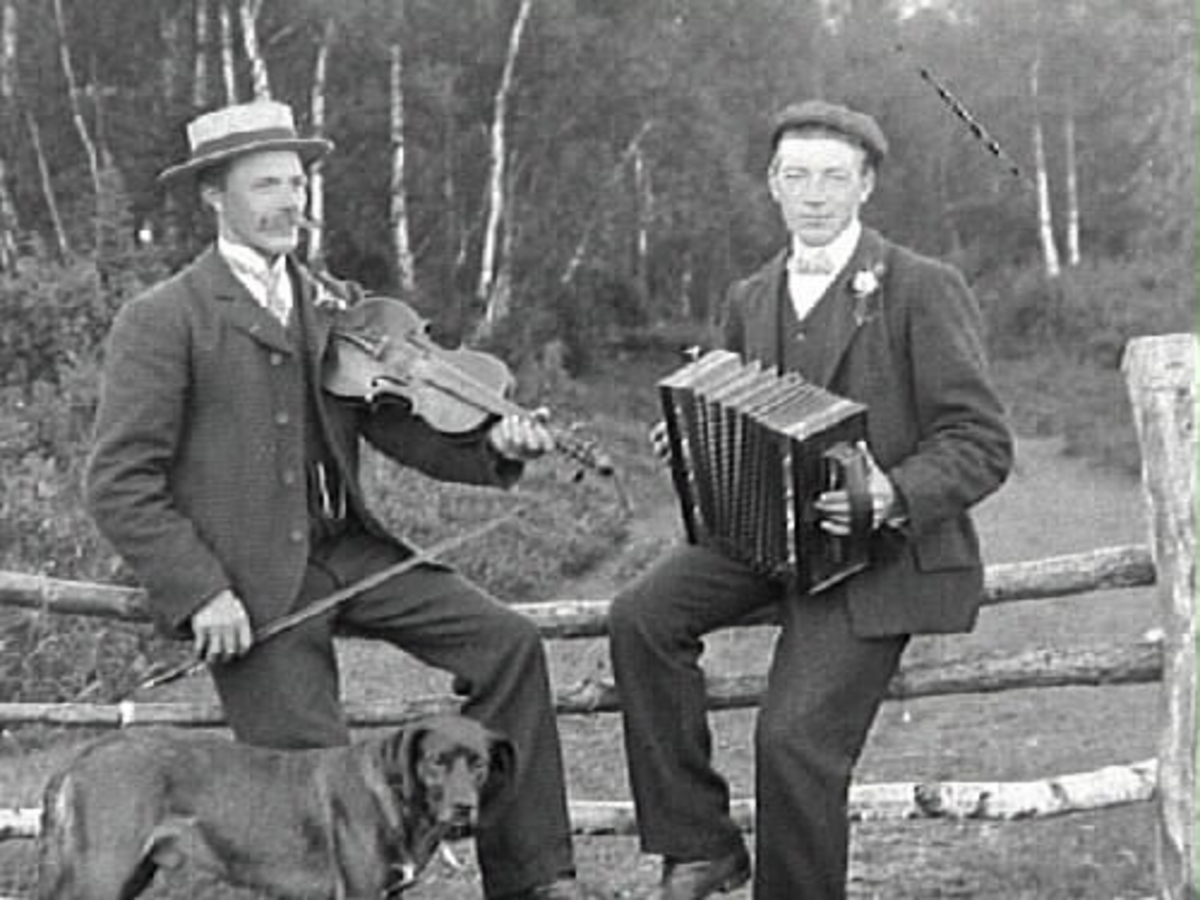 Edard Johansson med fiol, och brodern Klas Johansson med dragspel. Männen sitter på ett stängsel vid en hage med skog i bakgrunden. En hund står bredvid dem.