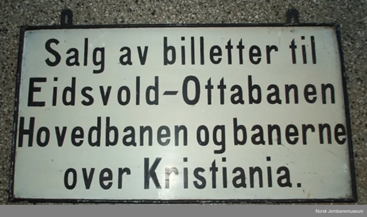Skilt tosidig "Salg av billetter til Eidsvold-Ottabanen Hovedbanen og banerne over Kristiania" og på baksiden "Salg av soveplads- og tilægsbilletter til hurtigtog"