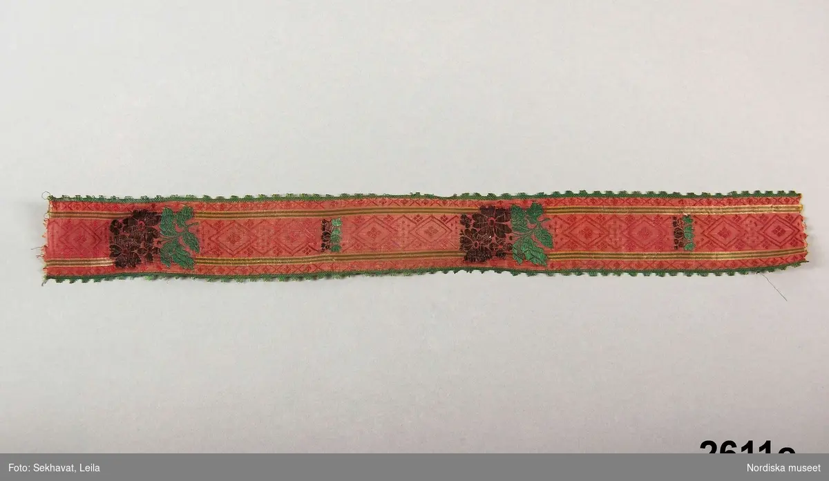 C.-D. Sidenband längd 38 cm, br. 4,5 cm, mönstervävda rosaröda med broscherade blommor i guld och grönt.

Hör till ungmorsdräkt med vithatt, i Delsbo kallad unggumdräkt, den som bars av bruden dagen efter bröllopet. Har tillhört säljaren fru Kihlström i Delsbo.
Plaggen 2603 - 2611 bildar en sådan dräkt men har tillhört olika personer, hopsamlad till en enhet av förmedlaren Lars Landgren.
/Berit Eldvik juni 2005