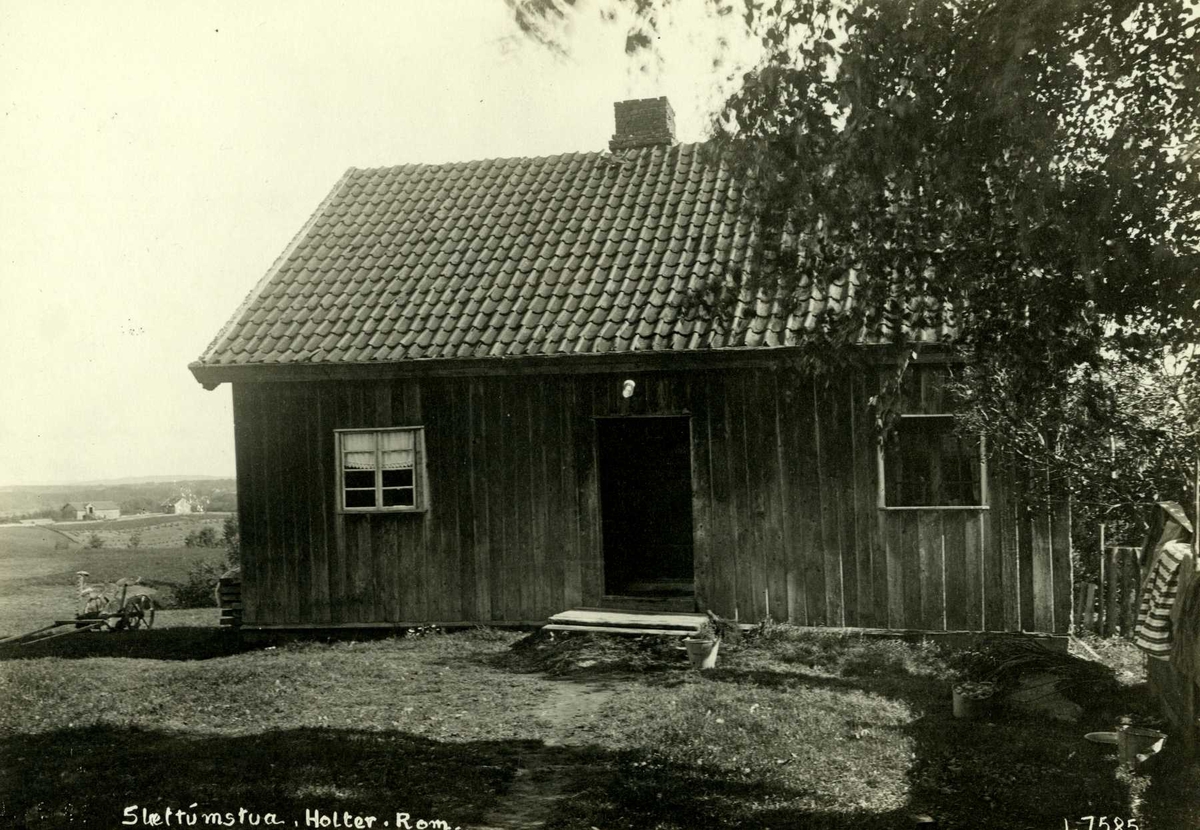 Slattumstuen, Nannestad, Øvre Romerike, Akershus. Lite våningshus sett mot gårdsplassen, hesteredskap ved siden av huset og utsikt mot bygda.