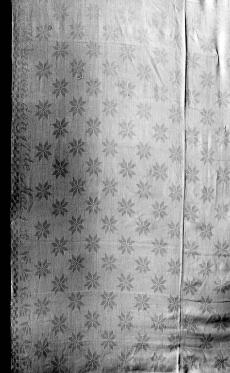 Hvit duk med stjernemønster, rankebord. Vevet i to lengder og sydd sammen. Fra 1836, linen er dyrket på Weisteen gård, Ringerike, Buskerud.  Norsk hjemmevevet damask.