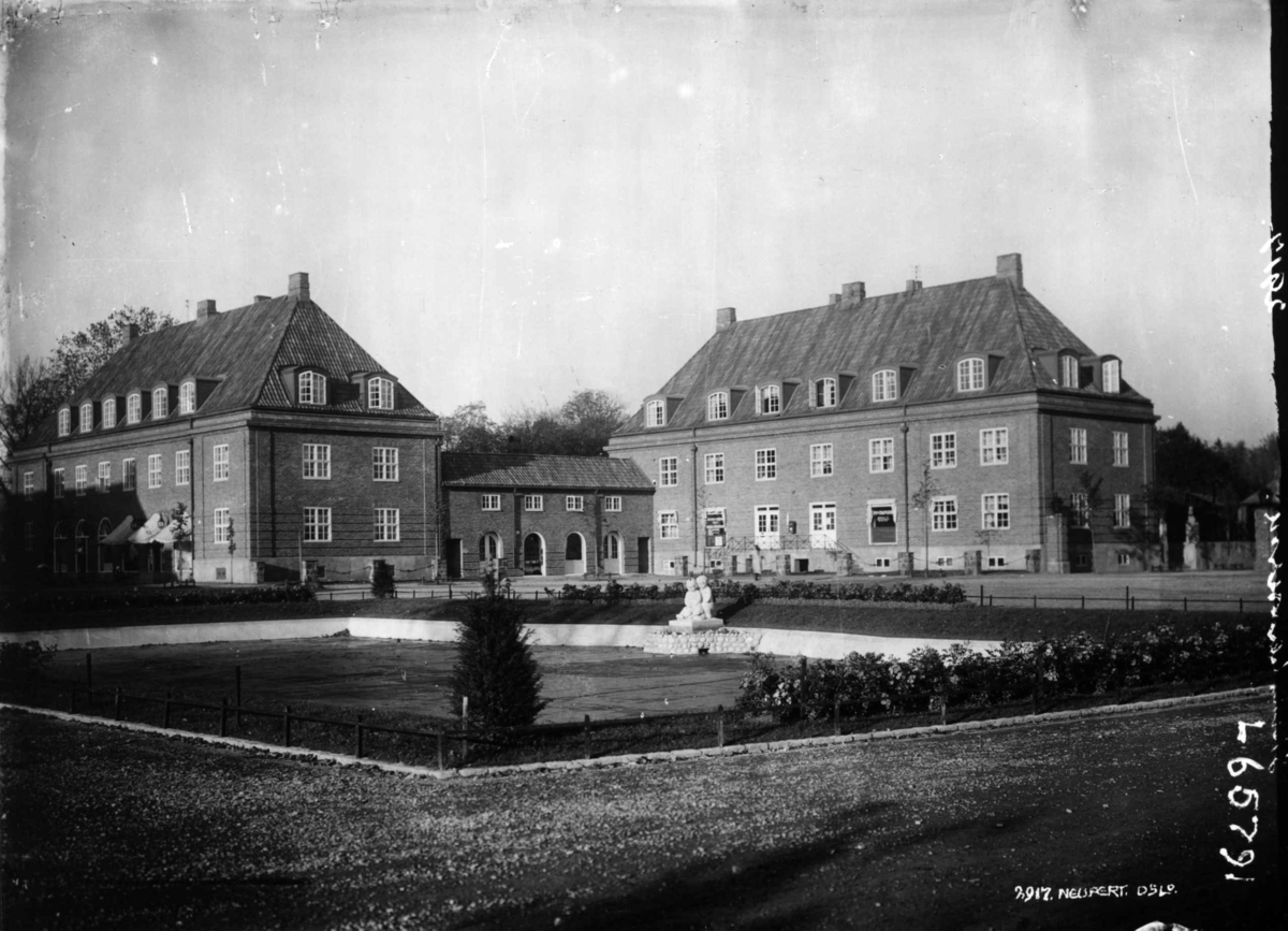 Damplassen, Ullevål Hageby, Oslo. 1925. Bebyggelse med forretninger i bakgrunnen. Beplantning i forgrunnen.