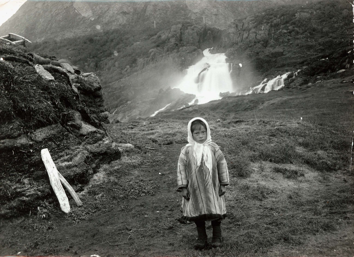 Pike i samisk søndagsdrakt, Valborg 8 år, oppstilt ved siden av gamme, hjemmet sitt, ved Adamsfjordfossen, Lebesby. 
Del av serie fra en forskningsreise i Øst-Finnmark 1909.