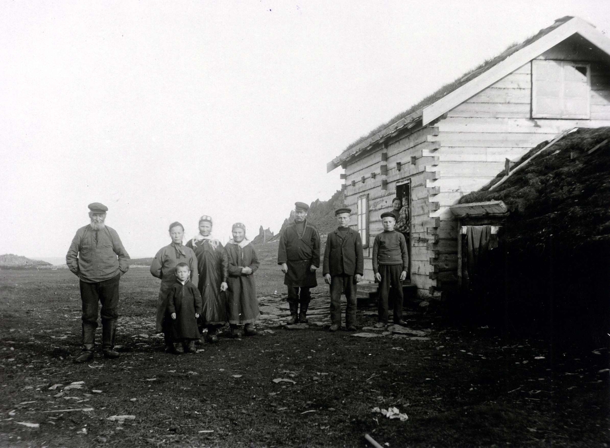 Persongruppe (10) utenfor bolig/gamme, Kongsfjord, Berlevåg, 5 i samisk drakt. Boligens hoveddel er laftet med jordgamme bygget inntil. 
Del av serie fra en forskningsreise i Øst-Finnmark 1909.