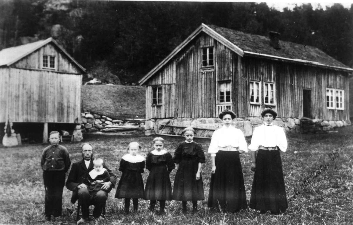 Butveit gård, Audnedal, Vest-Agder.
To kvinner , en mann og fem barn stilt opp for fotografering utendørs med gården som bakgrunn.